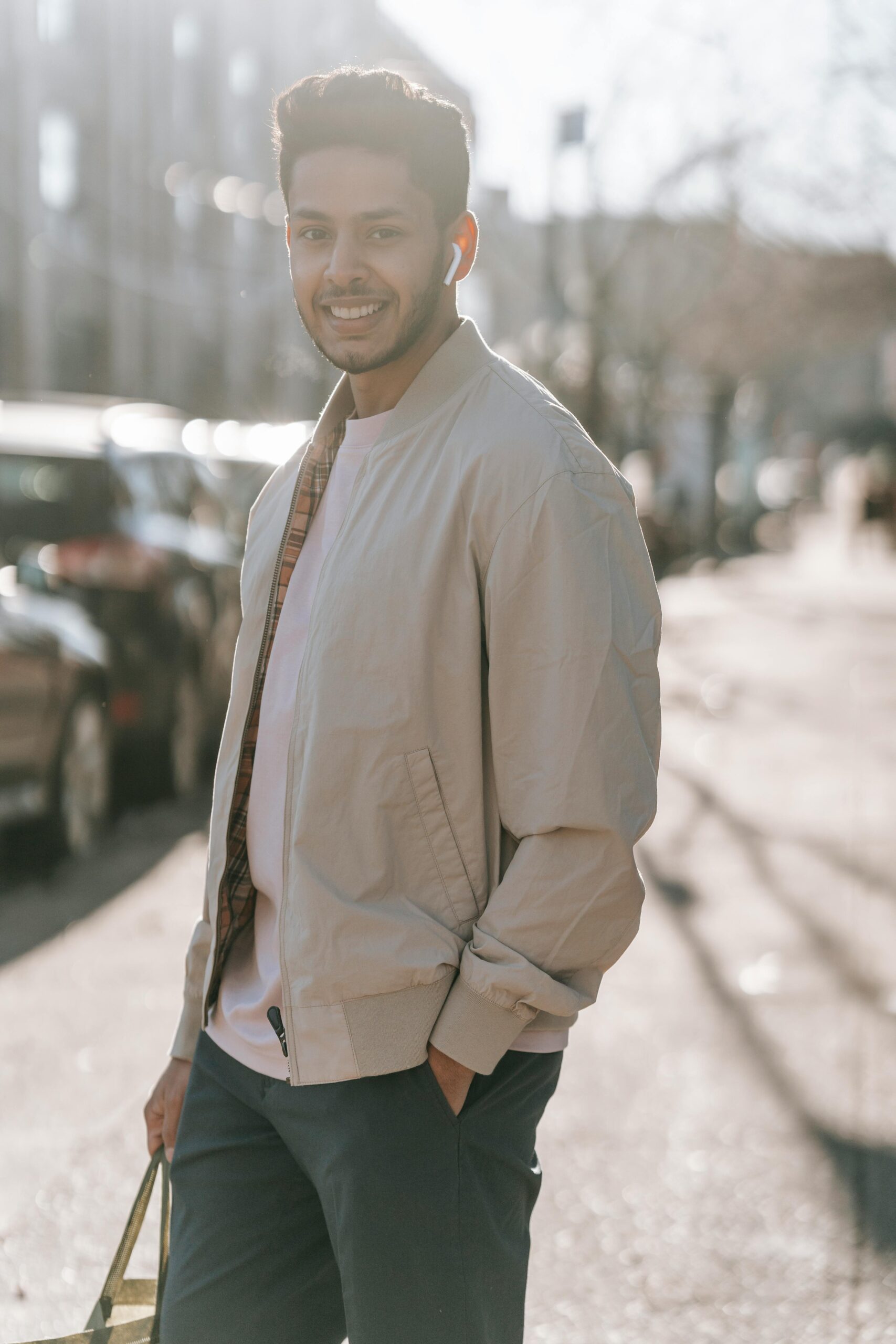 Cheerful ethnic male in trendy clothes with hand in pocket listening to music from earbud while looking at camera outdoors